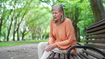 Elderly female with gray hair suffers from painful severe knee joint pain sitting on bench on street in urban city park. Upset woman massages leg muscles, could not walk, illness, cramps or rheumatism