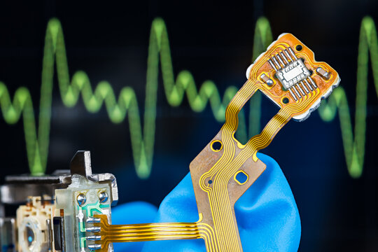 Integrated circuit of optical sensor, lens and flexible flat cable on technician hand in tech laboratory. Closeup dismantled CD-DVD disc drive pickup head on electronic oscilloscope screen background.