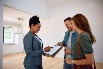 Happy couple analyzing housing plans with real estate agent while buying new home.