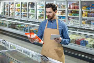 Male supermarket employee wearing apron uses tablet for inventory check while examining frozen food bags in grocery store. Focus on efficiency and accuracy in retail management.