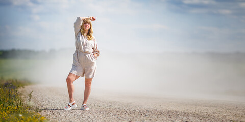 woman stands on dusty rural road under bright summer sky. She wears light linen romper and sandals,...