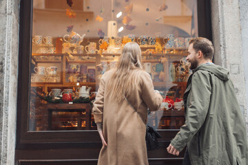 A couple admiring a shop window filled with colorful ceramic mugs and decorations. The woman has long hair and is wearing a beige coat, while the man is in a green jacket. The scene captures a cozy sh