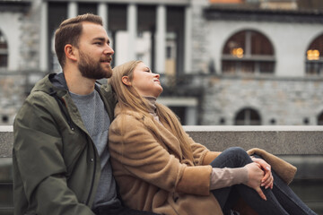Obraz premium A couple sitting together outdoors, enjoying a moment of relaxation. The man has a beard and is wearing a green jacket, while the woman is dressed in a beige coat, leaning back against him. They are i