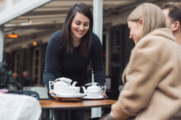 A waitress serving tea to two customers at an outdoor café. The waitress is smiling and holding a tray with teapots and cups. The customers are engaged in conversation, enjoying their time.