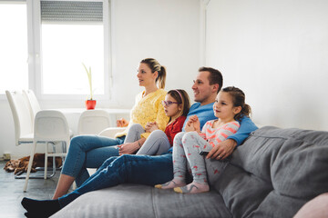 Obraz premium A family of four sitting together on a couch, watching TV. The mother and father are in casual clothing, with two young daughters beside them. The room has a bright, modern decor with a plant on the w