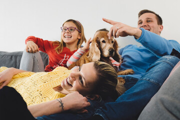 A family of four sitting together on a couch, watching TV. The mother and father are in casual...