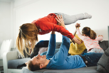A joyful father playing with his daughter on a couch, both laughing and having fun. The mother is in the background, relaxed. The scene captures a warm family moment.