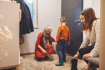 A woman in a red dress helps a young boy with his shoes in a cozy hallway. Another woman sits nearby, putting on her boots. Mother coming home from work, children hugging her with warm welcome.