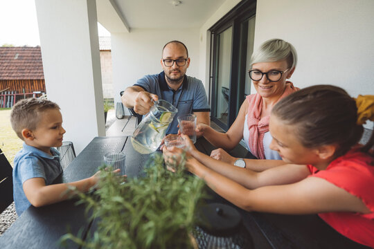 A woman serves a glass of water with lemon to a girl at a table, while a man sits nearby. They are enjoying a casual gathering outdoors, surrounded by plants and a cozy atmosphere.