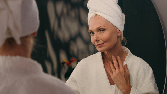 Middle-aged elderly old mature Caucasian 50s woman smile looking in mirror reflection touching face check facial wrinkles clean hydrated skin anti aging cosmetic beauty skincare treatment in bathroom