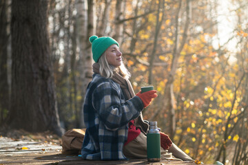 Pleased thoughtful woman rest alone walking in fall forest, drinking hot tea from thermos sitting on wooden path. Satisfied relaxed blonde female in warm clothes enjoys stroll in autumn woodland park