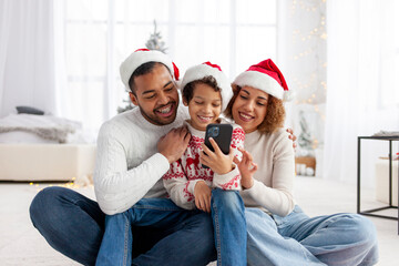 african american family in santa hat hugging at home for christmas and giving smartphone to son, teenage boy celebrating new year with mom and dad and using phone, parents choosing online and typing