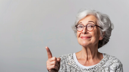 A confident elderly woman with gray hair and glasses, wearing a cozy gray sweater, smiling gently and pointing upward with a thoughtful expression, set against a clean white background