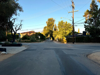 typical American neighborhood empty residential street intersection (driveways, homes, parked cars, telephone poles and wires, signs trees) beneath a bright blue summer sky in Los Angeles, California