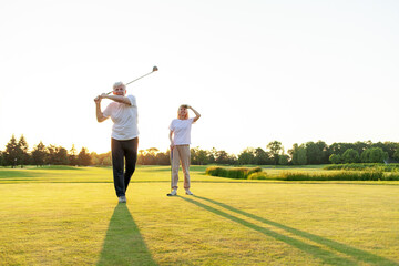 elderly senior couple in uniform playing golf on golf course at sunset, old man swinging club at ball outdoors, woman looking ahead into distance