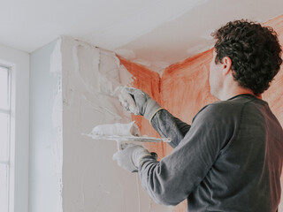 A young caucasian guy applies fresh putty with a small spatula on the wall near the window