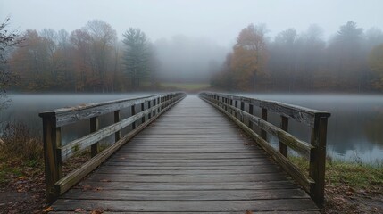 Naklejka premium A serene wooden bridge extends into a foggy landscape, surrounded by autumn foliage.