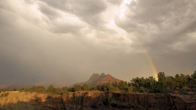 A horizontal bolt of lightning flashes across the sky above Smithsonian Butte in Southern Utah, USA below grey clouds while a rainbow arcs from the ground up to the sky seen from Gooseberry Mesa.