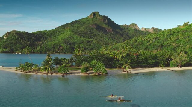 Two people paddling an outrigger canoe near tropical island with white sand beach, green towering mountains in background. Active sports lifestyle. French Polynesia. remote wild nature paradise aerial
