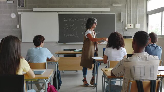 Mature female teacher handing out exams to high school students in classroom. Education lifestyle concept.