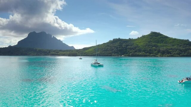 Sailboats anchored in turquoise lagoon, green mountains tropical island in background. Crystal clear water reflects blue sky. Yacht catamaran boat tour to French Polynesia. Exotic summer luxury travel