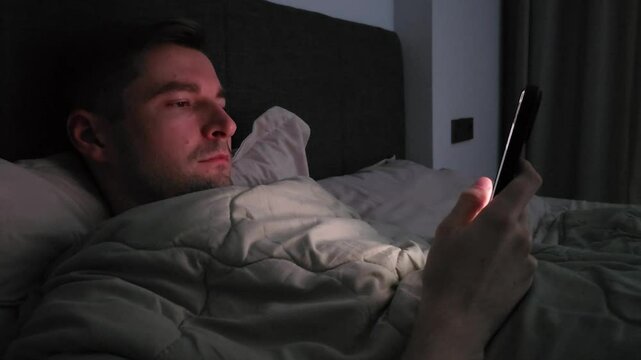 Young white man using smartphone while lying in bed at night, having sore and tired eyes. No sleep from insomnia