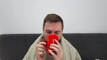 Man with irritated red nose blowing into tissue, holding wipes, and drinking hot tea or coffee from a red cup. Wrapped in a blanket on a couch. Health and illness concept. Design for poster, banner, a