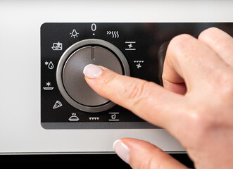 Woman adjusting modern white oven in kitchen.  Female hand pushing and turning knobs on oven control panel