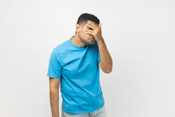 Portrait of curious attractive unshaven man wearing blue T- shirt standing looking at camera through fingers, peeking, sees something shameful. Indoor studio shot isolated on gray background.