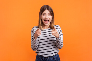 Fototapeta premium Portrait of excited amazed woman with brown hair pointing at camera choosing you, screaming with happiness, wearing striped shirt. Indoor studio shot isolated on orange background