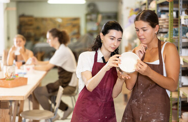 Interested young girls wearing aprons viewing attentively handmade clay bowl in pottery workshop - Powered by Adobe