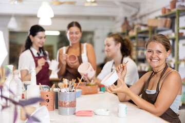 Interested young woman in pinafore drawing on handmade clay jar with paintbrush in art studio