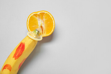 Fresh banana in condom with lipstick kiss and orange on grey background, closeup. Sex education...