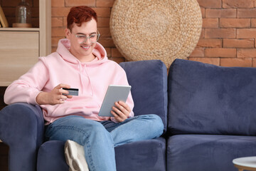 Handsome young man with credit card and tablet computer sitting on sofa while shopping online