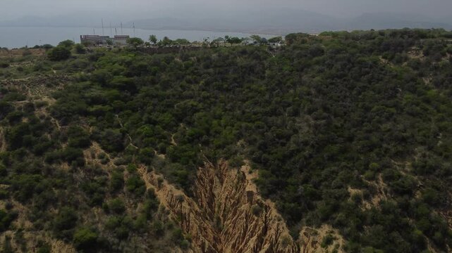 Aerial tracking by drone of a compact blue car, rolling on the ridge of a mountain, with arid vegetation and beach in the background