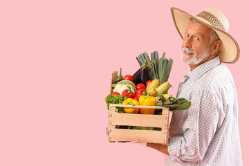 Happy mature male farmer with wooden box full of different ripe vegetables on pink background