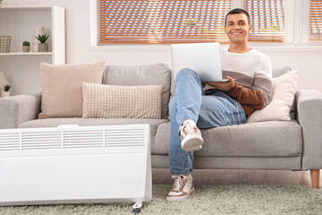 Young man with laptop warming near radiator at home