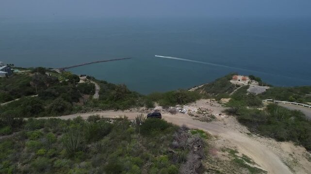 Aerial tracking by drone of a compact blue car, rolling on the ridge of a mountain, with arid vegetation and beach in the background