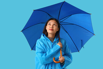 Thoughtful young Asian woman in raincoat with umbrella on blue background