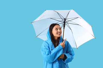 Beautiful young Asian woman in raincoat pointing at umbrella on blue background