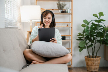 A young woman sitting on a sofa, using a tablet and wearing headphones in a cozy, modern living room with stylish decor.