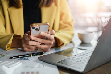 Fototapeta premium Professional woman in yellow blazer using smartphone at work desk with laptop, documents, and coffee cup in a modern office.