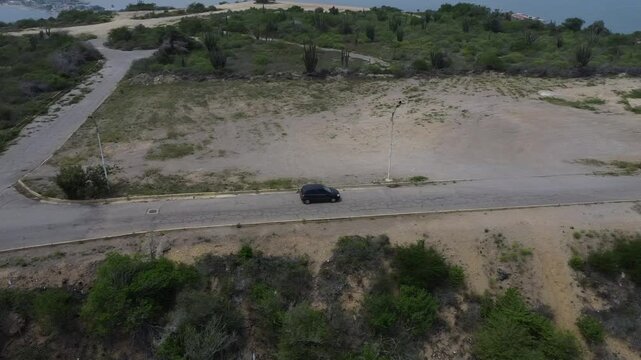 Aerial tracking by drone of a compact blue car, rolling on the ridge of a mountain, with arid vegetation and beach in the background