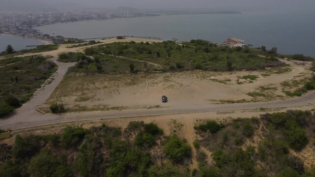 Aerial tracking by drone of a compact blue car, rolling on the ridge of a mountain, with arid vegetation and beach in the background