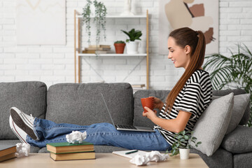 Obraz premium Beautiful female writer with laptop and cup of tea sitting on sofa at home