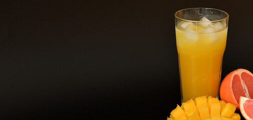 A tall glass of a mixture of fruit juices with ice on a black background, with pieces of ripe mango and grapefruit in it.