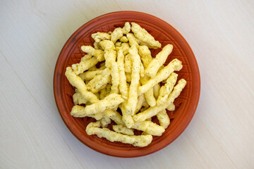 Tasty corn stick chips in a clay plate on a light wooden surface. Kurkure American-style cream and onion flavor chips, a delicious savory snack food. Top view.