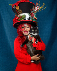 Woman with Christmas makeup, wearing huge hat with Christmas ornaments, hugging her black and white cat.
