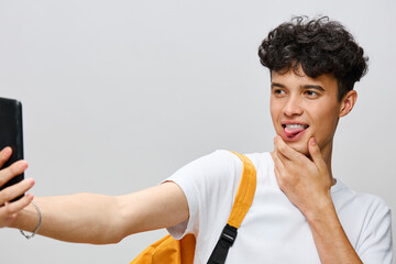Young man taking a selfie with a playful expression against a light gray background, wearing a...