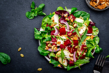 Vegan salad with baked beetroot, fresh pear, arugula, lettuce, red onion, chard, pumpkin seeds and walnut, black table. Top view